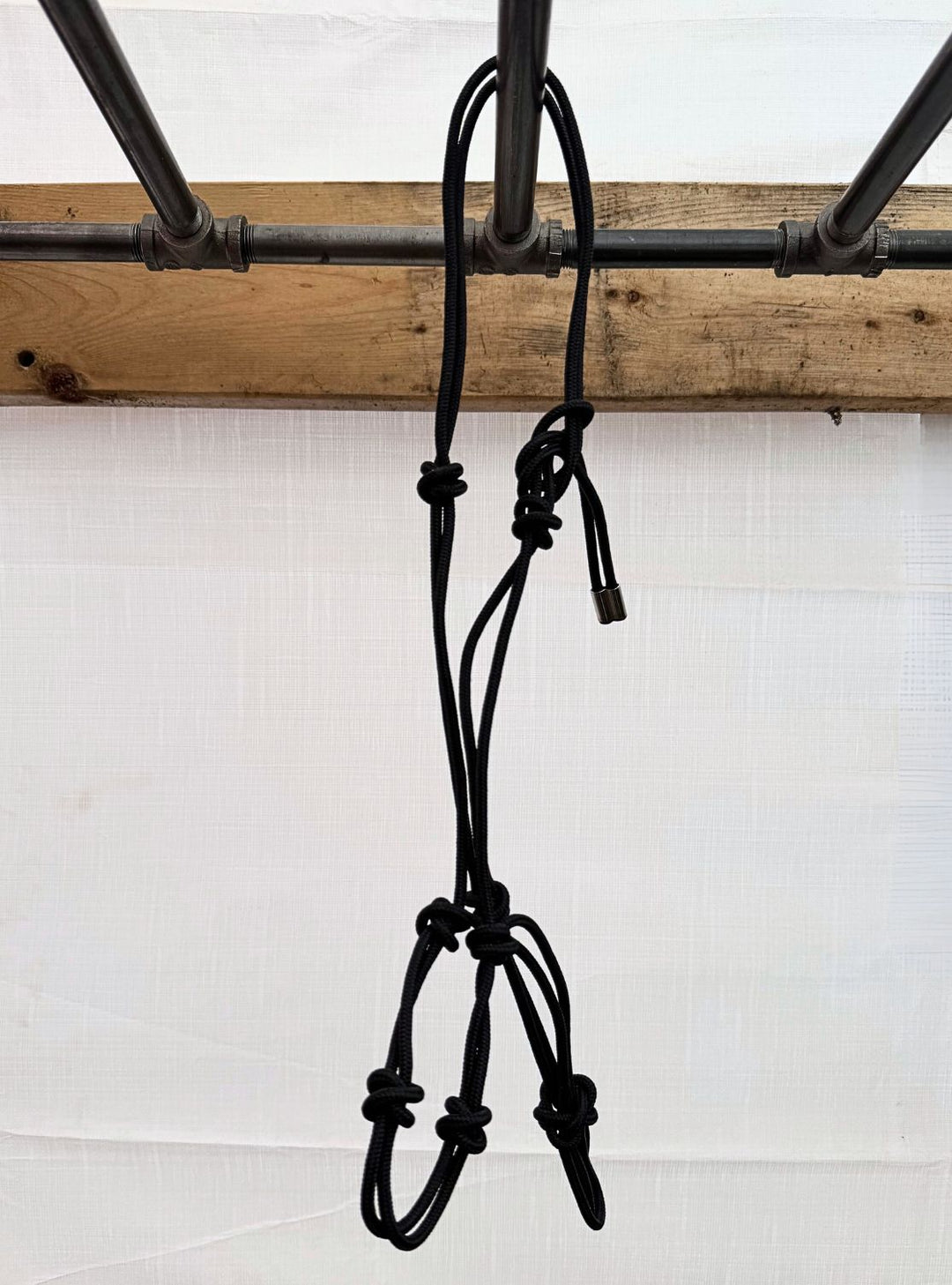 A sturdy black rope halter with multiple knots hanging from a metal post backed by a wooden plank.